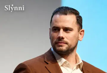 Grzegorz Collins, znany z programów telewizyjnych, w eleganckim brązowym garniturze. Jego wiek jest trudny do określenia, ale prezentuje się dojrzale.