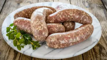 Kilka surowych kiełbasek białych parzonych ułożonych na marmurowej desce, ozdobione gałązką zielonych liści.