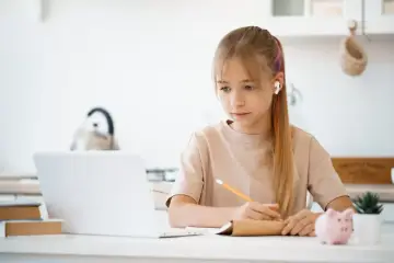 Dziewczynka z słuchawkami w uszach uczy się przy laptopie, notując w zeszycie. Tak wygląda nauka w chmurze.