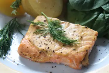 Pieczony łosoś w sosie z koperkiem, obok ziemniaków i zielonych liści.