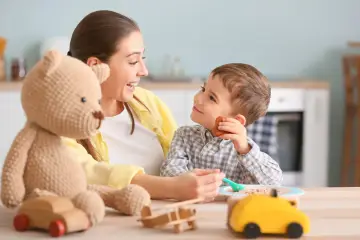 Uśmiechnięta opiekunka karmi chłopca. Na stole pluszowy miś i zabawki. Zastanawiasz się, ile opiekunki biorą za godzinę?