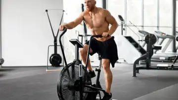 Mężczyzna z brodą intensywnie ćwiczy cardio na rowerze stacjonarnym w siłowni.