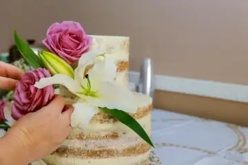 Ręce dekorują dwupiętrowy tort nagus, dodając róże i lilie. Piękny sposób, jak ozdobić tort kwiatami.