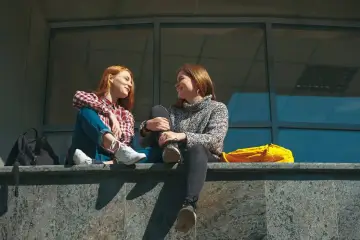 Dwie dziewczyny rozmawiają, siedząc na murku. Jedna z nich ma rude włosy i kratę. Czy to rozmowa o tym, czym jest absencja?