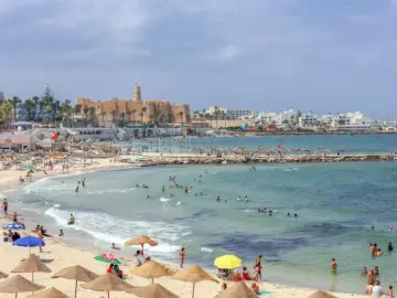 Tłum ludzi cieszy się słońcem i morzem na plaży w Tunezji, z widocznym w oddali zabytkowym fortem.
