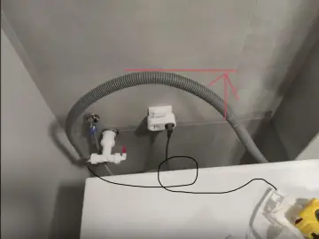 Szara rura odpływowa pralki w ścianie, podłączona do białego syfonu. Obok gniazdko elektryczne.