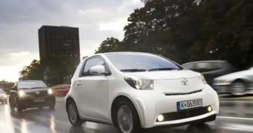 Biały Toyota iQ jedzie po mokrej ulicy, mijając inne samochody. W tle widać budynek i drzewa.