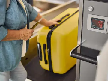 Kobieta z żółtą walizką i paszportem przy wadze bagażowej. Pamiętaj, czego nie można przewozić w bagażu podręcznym, by uniknąć problemów.