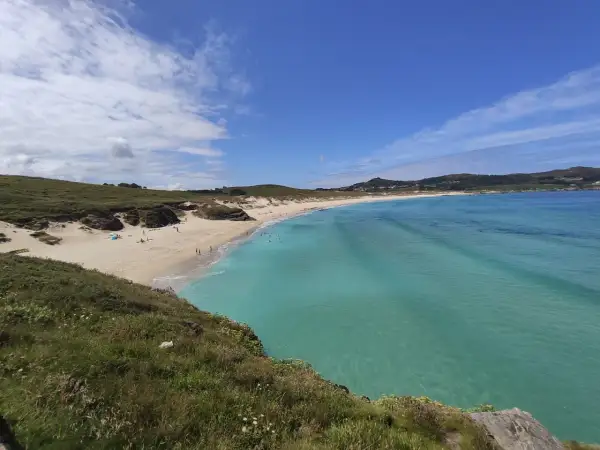 Vista panorámica de la playa de Santa Comba, con arena dorada, aguas turquesas y colinas verdes. Algunas personas disfrutan del sol y el mar.
