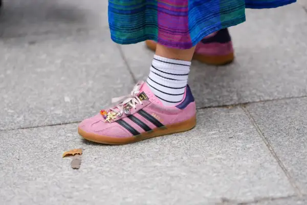 Różowe buty sportowe Adidas Gazelle z czarnymi paskami i charmsami, białe skarpetki w paski. W tle widać drugie, podobne buty w innym kolorze.