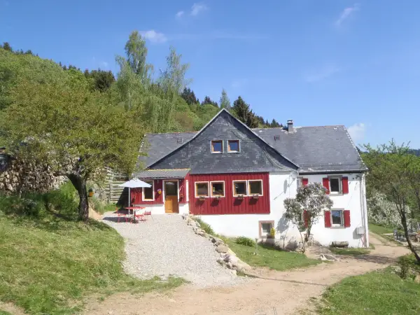 Maison de campagne avec terrasse et parasol, idéale pour un camping à la ferme dans les Vosges. Le soleil brille sur ce paysage verdoyant.