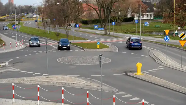 Samochody zbliżają się do ronda. Widoczny jest kierunkowskaz na rondzie, a także znaki drogowe i bariery.