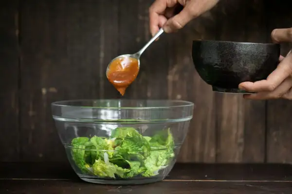 Ein Löffel mit braunem Salatdressing tropft in eine Schüssel mit knackigem Salat.