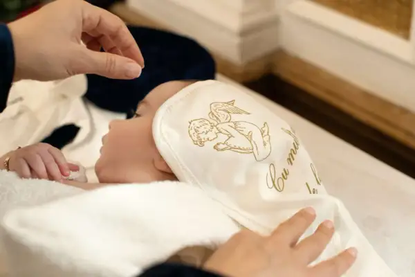 Dziecko otulone w biały kocyk z aniołkiem, przygotowywane do chrztu. To ważny moment, który potwierdzi zaświadczenie od księdza do chrztu.