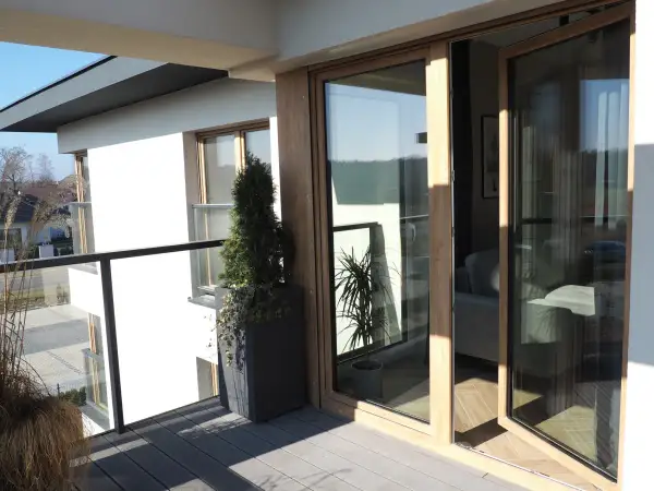 Nowoczesne okna dzielone z drewnianymi ramami otwierają widok na balkon z roślinnością i wnętrze domu.