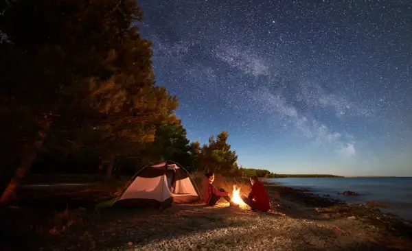 Nocne biwakowanie pod rozgwieżdżonym niebem. Dwoje ludzi siedzi przy ognisku obok namiotu z materiału namiotowego, na kamienistej plaży.