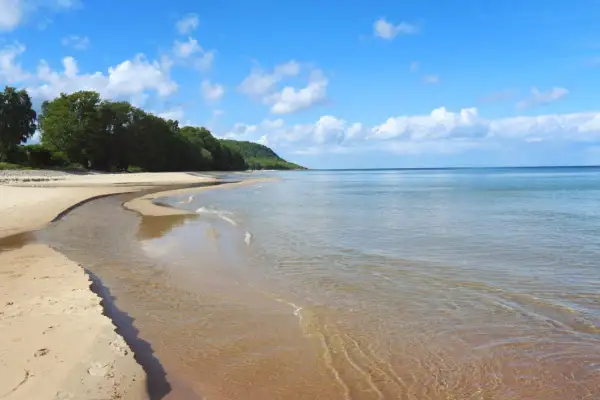En stilla stenshuvud strand med klart vatten som möter en sandig kustlinje. Grönska och kullar syns i bakgrunden under en blå himmel med vita moln.