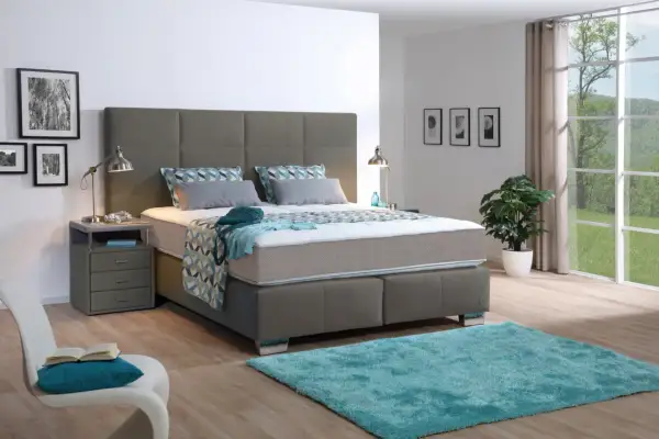 Ein modernes Schlafzimmer mit einem grauen Boxspringbett, das mit einer geometrisch gemusterten Decke und Kissen bezogen ist. Ein türkiser Teppich liegt davor.
