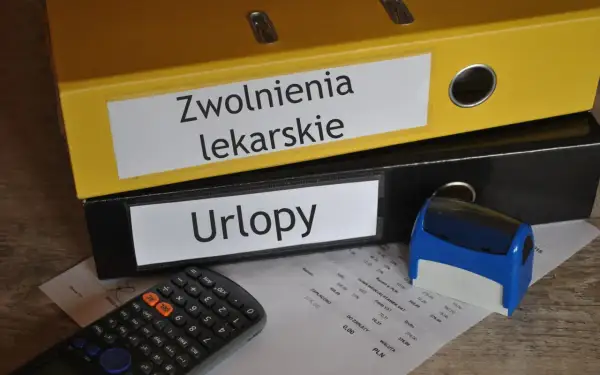 Zółty segregator "Zwolnienia lekarskie" i czarny "Urlopy" obok kalkulatora i pieczątki. Dokumenty na biurku sugerują, że kod a na zwolnieniu jest w trakcie rozliczeń.