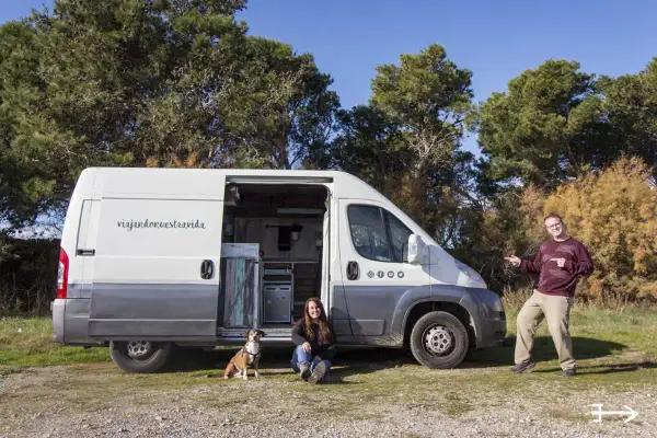 Pareja y perro posan junto a su furgón vivienda, listos para viajar.