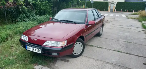 Czerwony Daewoo Espero z tablicą rejestracyjną WN-3841R zaparkowany przy zarośniętym poboczu i betonowej drodze.