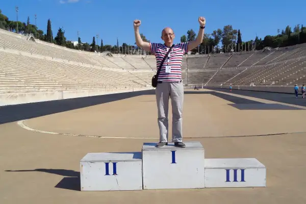 Mężczyzna na podium w historycznym stadionie panatenajskim, z rękami w górze, świętuje zwycięstwo.