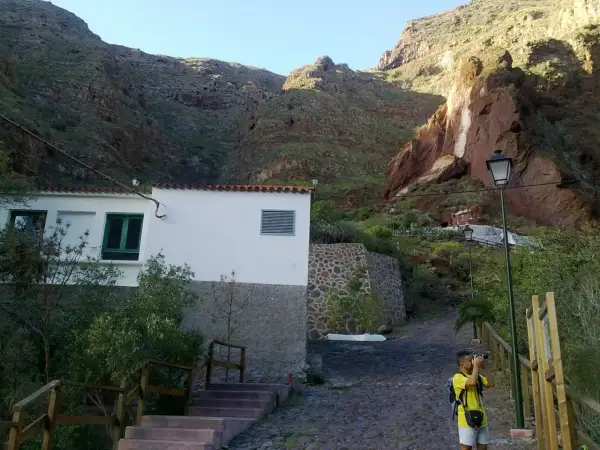 Un niño toma fotos en un camino empedrado, con una casa blanca y montañas rojas de fondo, cerca de la cueva bermeja.