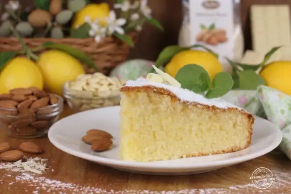 Una fetta di soffice torta caprese al limone, guarnita con zucchero a velo e foglioline di menta, circondata da limoni freschi e mandorle.
