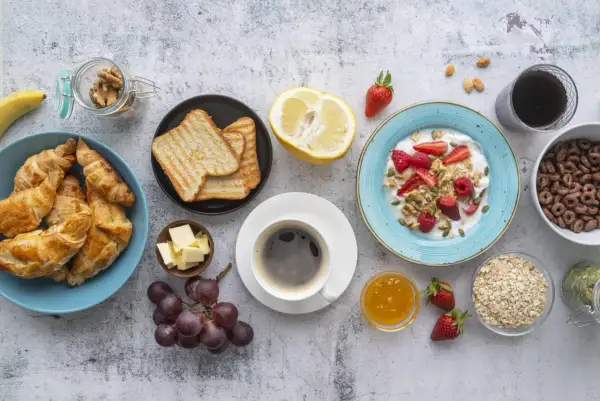 Un tavolo pieno di idee su cosa mangiare a colazione: croissant, toast, yogurt con frutta, caffè, uva e cereali.