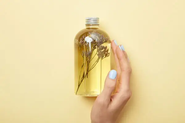 Eine Hand mit hellblauen Nägeln berührt eine Flasche mit gelbem Öl und getrockneten Blumen. Perfekt für alle, die Duschgel selber machen wollen.