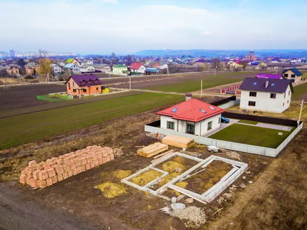 Budowa domku na działkę zrób to sam. Widać fundamenty, sterty cegieł i drewno budowlane. W tle ukończone domy.