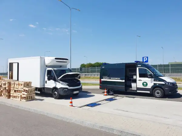 Kontrola drogowa: biała ciężarówka z otwartą maską i stosy palet obok, obok zielony bus Inspekcji Drogowej. Sprawdzana jest ładowność w dowodzie rejestracyjnym.