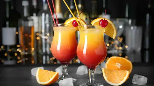 Dwa kolorowe drinki z wódką, ozdobione pomarańczą i wiśnią, na tle baru.