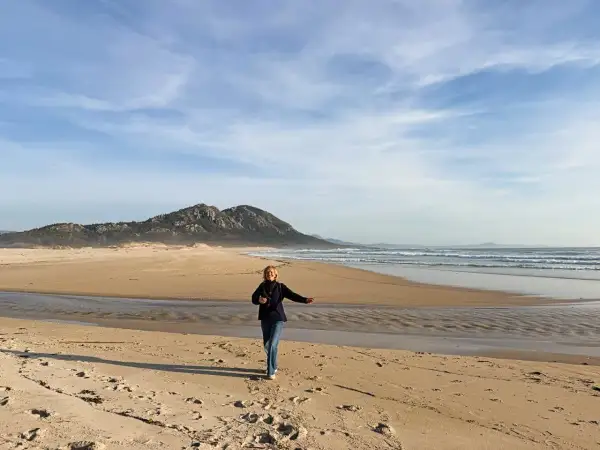 Mujer corre en la playa de Muros, Galicia, con montañas al fondo y el mar azul.