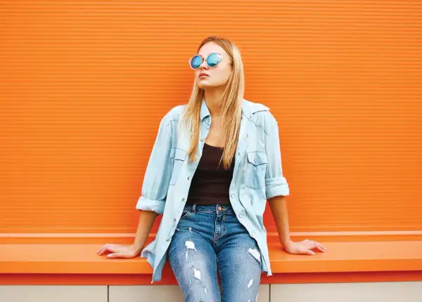 Blondynka w okularach i jeansach, z rozpiętą błękitną koszulą. Idealny przykład, jak nosić koszulę do jeansów na luzie.