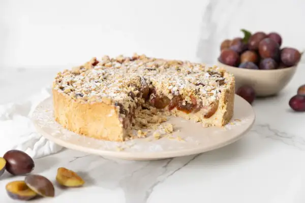 Ein köstlicher Zwetschgenkuchen mit Streuseln, bestäubt mit Puderzucker, steht auf einem Teller. Daneben liegen frische Zwetschgen.