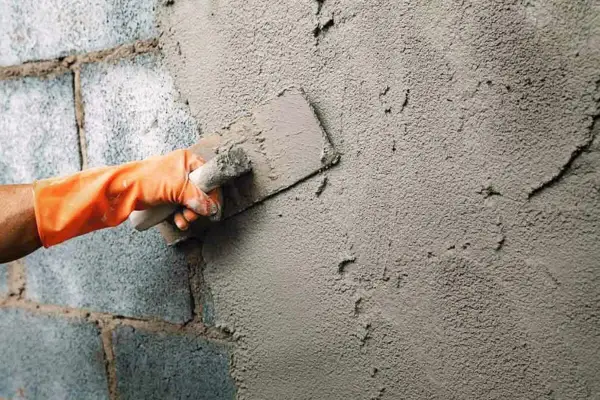 Mano con guante naranja aplica enfoscado rugoso a pared de bloques de cemento con llana.