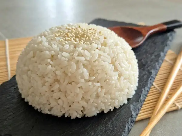 Ein Berg aus weißem Reis, bestreut mit Sesam, auf einem schwarzen Stein. Daneben liegen Stäbchen und ein Holzlöffel. Das ist die Basis, was bedeutet Sushi.
