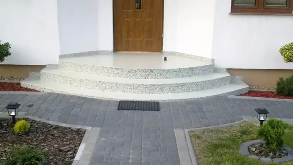 Nowe schody z żywicy na balkon, z kamyczkami, prowadzące do drewnianych drzwi. Wokół kostka brukowa i roślinność.