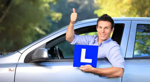 Hombre sonriente en coche con cartel "L", pulgar arriba. Pregunta: ¿cuanto tiempo hay que llevar la L?