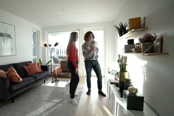 Två personer i ett ljust vardagsrum med stilren inredning. En mörk soffa med kuddar, ett vitt soffbord och en golvlampa. Hyllor med växter och dekorationer.
