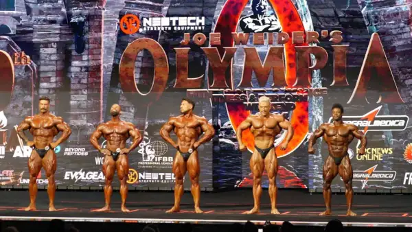 Fünf muskulöse Männer posieren auf der Bühne bei einem Bodybuilding-Wettkampf. Die "Joe Weider's Olympia" Bühne ist im Hintergrund zu sehen.