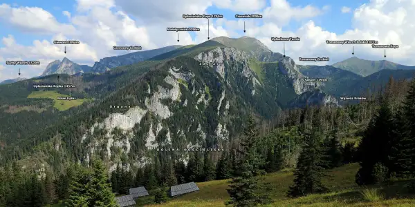 Widok na Tatry z Doliną Kościeliską. W oddali szczyty: Giewont, Ciemniak, Tomanowy Wierch Polski. Na pierwszym planie widać drewniane hale stoły.