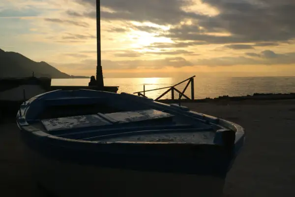 Un'imbarcazione blu riposa sulla riva al tramonto, con il sole che filtra tra le nuvole e illumina il mare calmo.