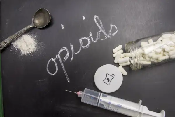 Napis "opioidy" na tablicy, obok łyżeczki z proszkiem, strzykawki, kapsułek i symbolu recepty.