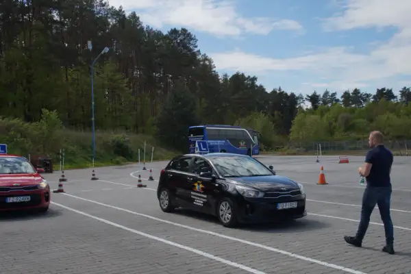 Na placu manewrowym, gdzie obowiązuje ruch lewostronny, czarny samochód szkoleniowy i czerwony pojazd czekają na kursantów. W tle niebieski autokar.