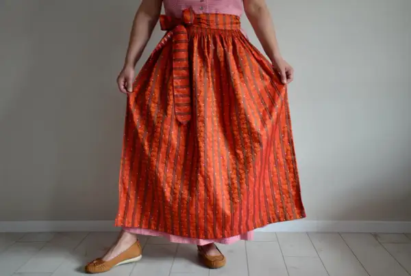 Eine Frau trägt ein Dirndl mit einer leuchtend orangefarbenen Dirndlschürze, die mit Streifen und kleinen Tupfen gemustert ist.