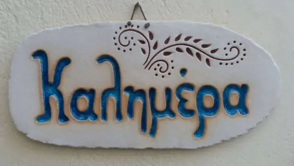 Ceramiczna tabliczka z napisem "Καλημέρα" (Dzień dobry) po grecku, ozdobiona roślinnym motywem.