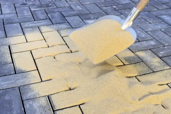 Schaufel mit Sand zum Pflastersteine verfugen. Sand rieselt auf dunkle Steine, um Fugen zu füllen.