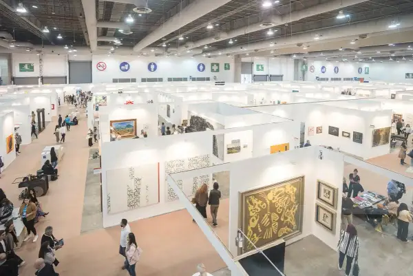 A bustling scene at the Mexico City Art Fair, with visitors exploring numerous white-walled booths showcasing diverse artworks.